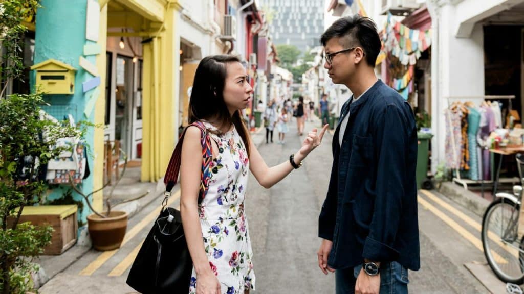 A woman points at a man while standing with him on a narrow street.
