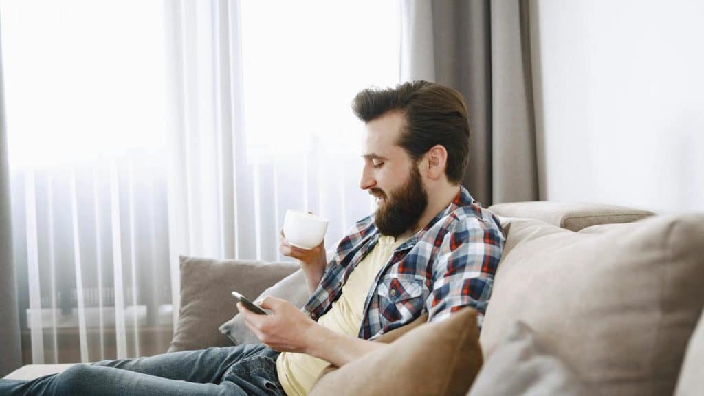 A bearded man sitting on a couch using his phone and holding a cup.