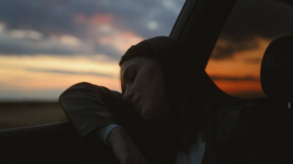 A woman resting her head against a car window at sunset.