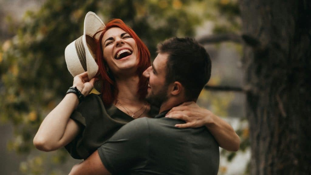 A man lifts a laughing woman with red hair who is holding a white hat.