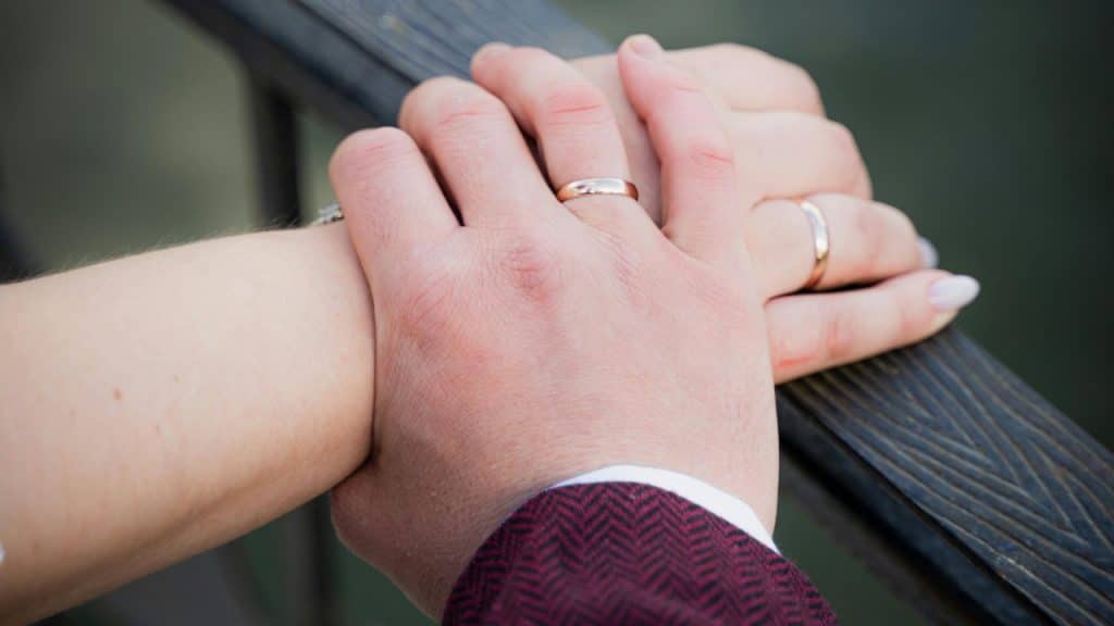 Two hands wearing wedding rings are gently clasped together on a railing.