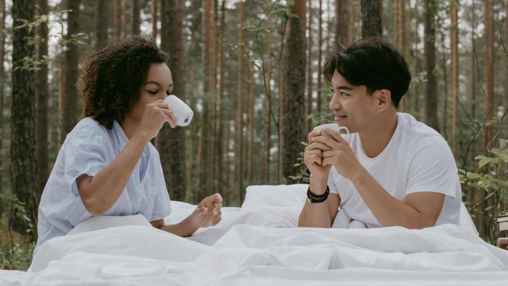 A couple sitting in bed outdoors drinking coffee.