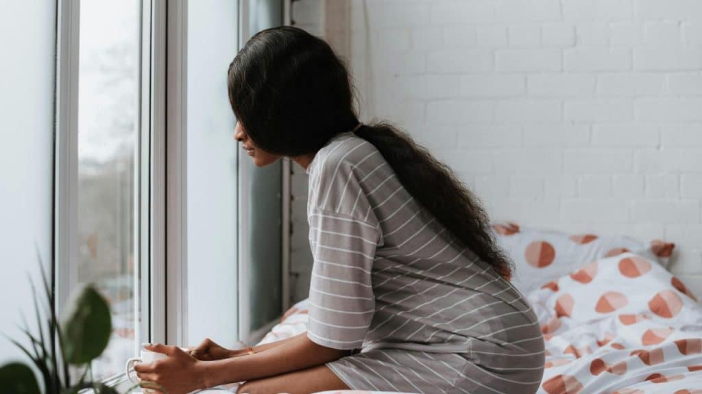 A woman sitting on a bed by a window, looking outside thoughtfully.