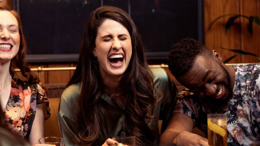 Three people laugh together while sitting at a table with drinks in a dimly lit room.