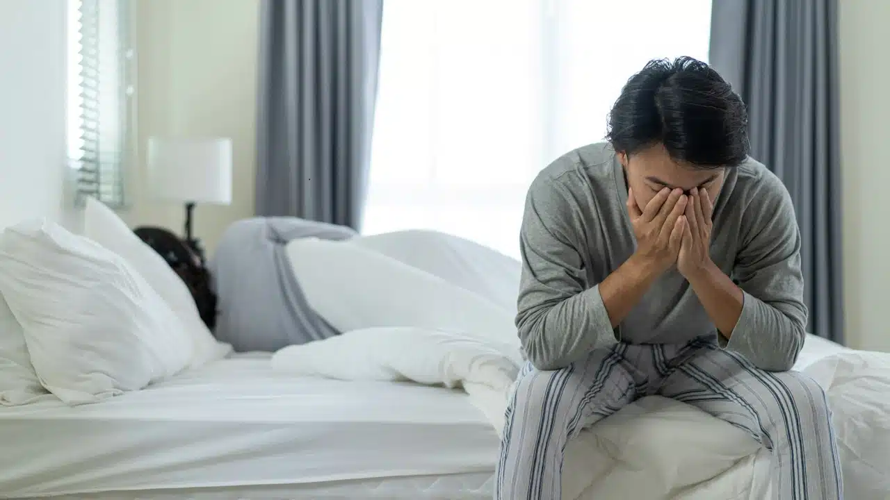 A man covering his face with his hands is sitting on one side of the bed while his wife is sleeping on the other side of the bed.