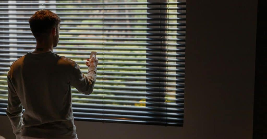 A man standing by window blinds while checking his phone.