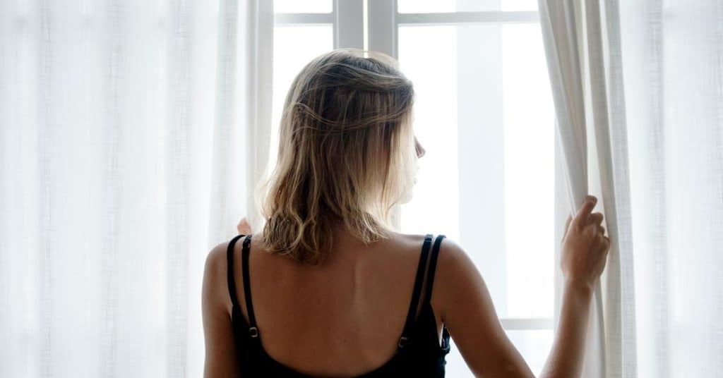A woman seen from behind, standing at a window and holding open sheer curtains.