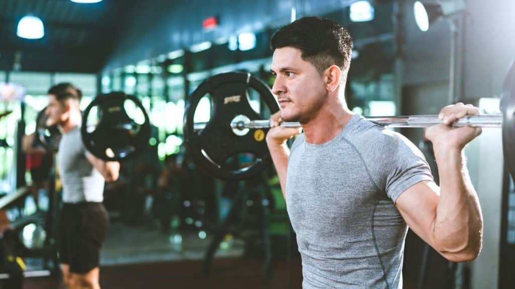 A man in a grey athletic shirt performs squats with a weighted barbell in a gym.