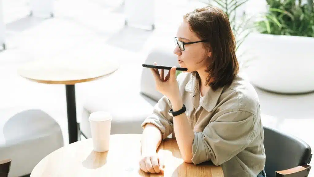 A woman sitting at a table in a café using her smartphone’s voice assistant.
