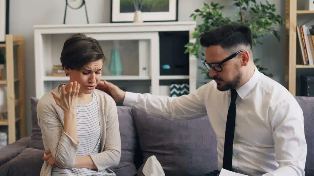 A man in a white shirt and tie places a hand on a crying woman’s shoulder.