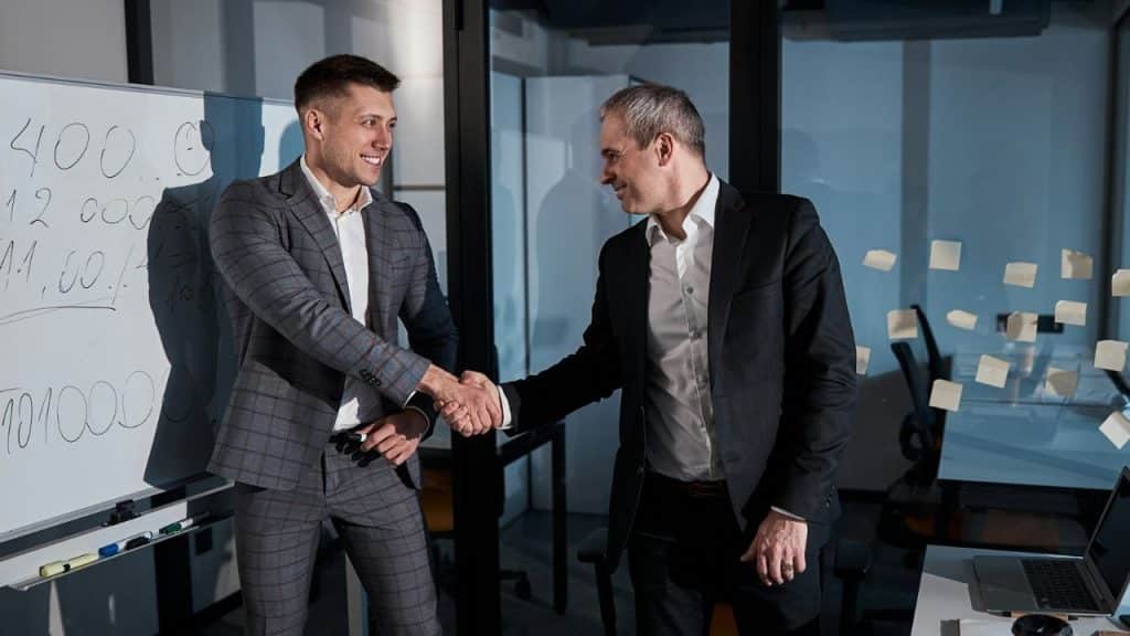 Two men in suits shake hands and smile in an office beside a whiteboard.