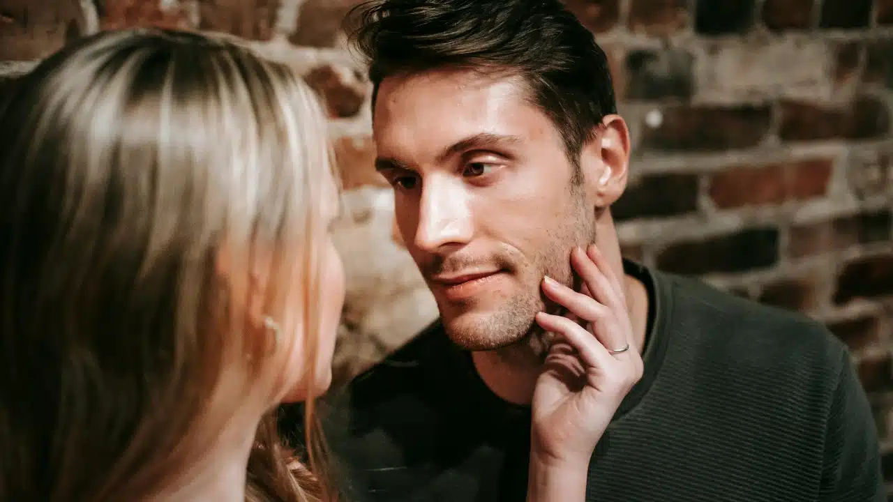 A woman’s hand rests on a man’s face while they look at each other closely.