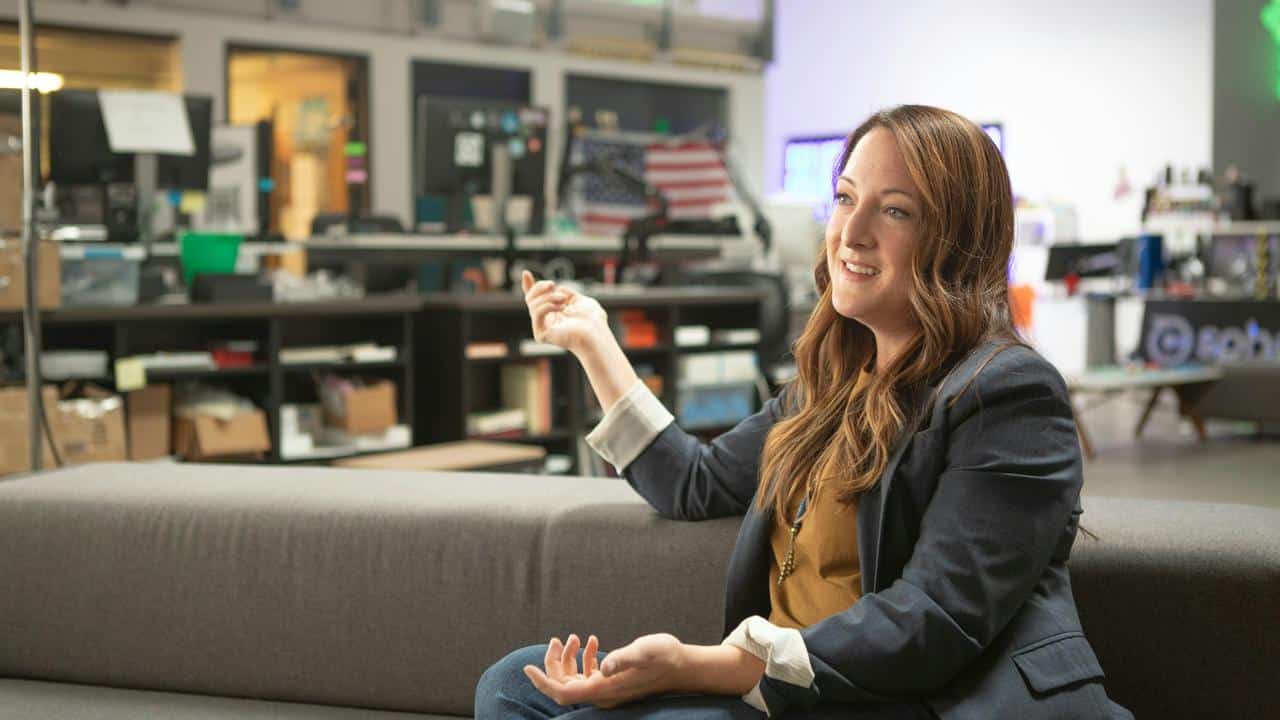 A woman sitting on a couch in an office, smiling while gesturing during a conversation.