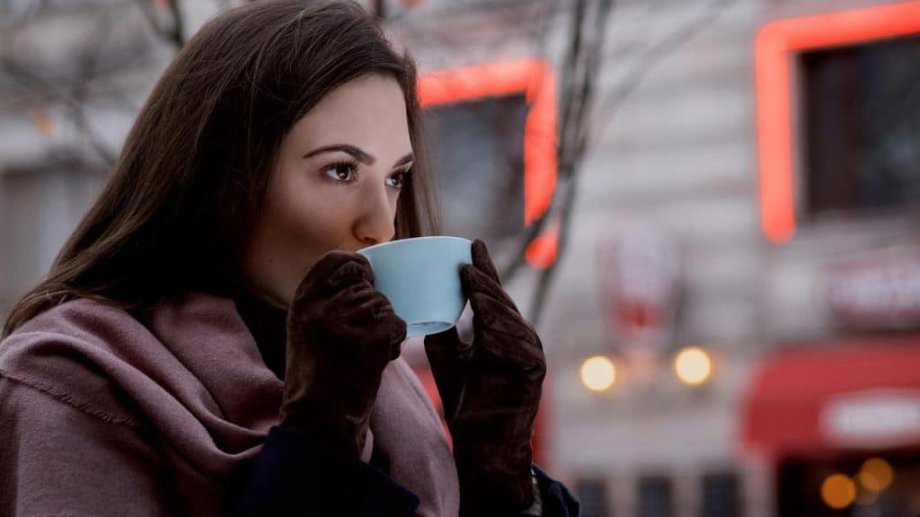 A woman wearing gloves drinks from a cup outdoors in a city setting.