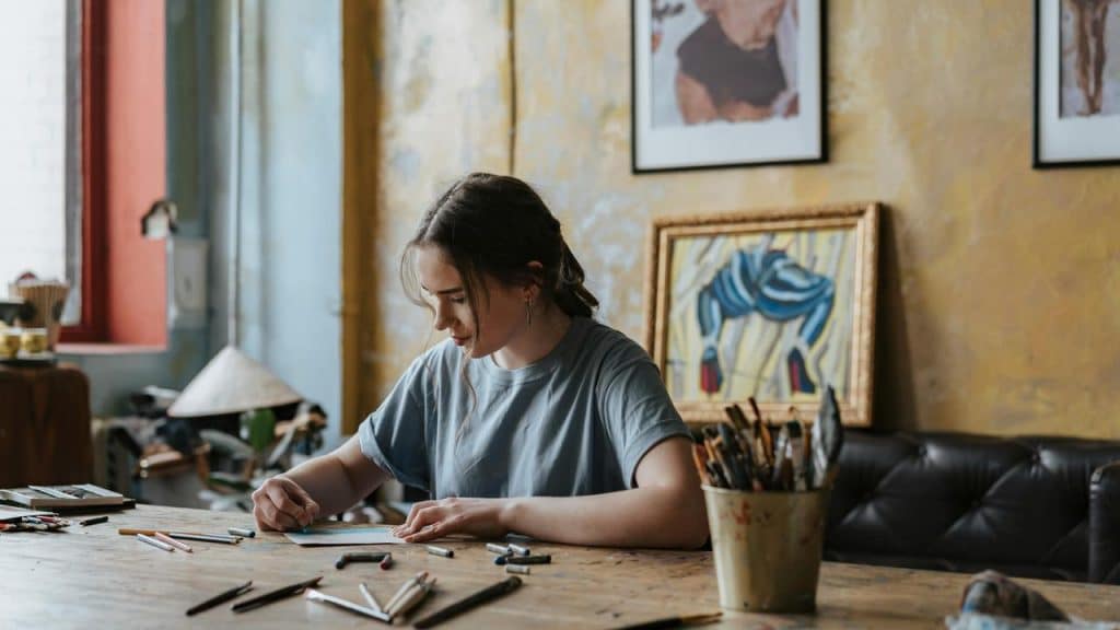 A young woman drawing at a cluttered desk in an art studio.