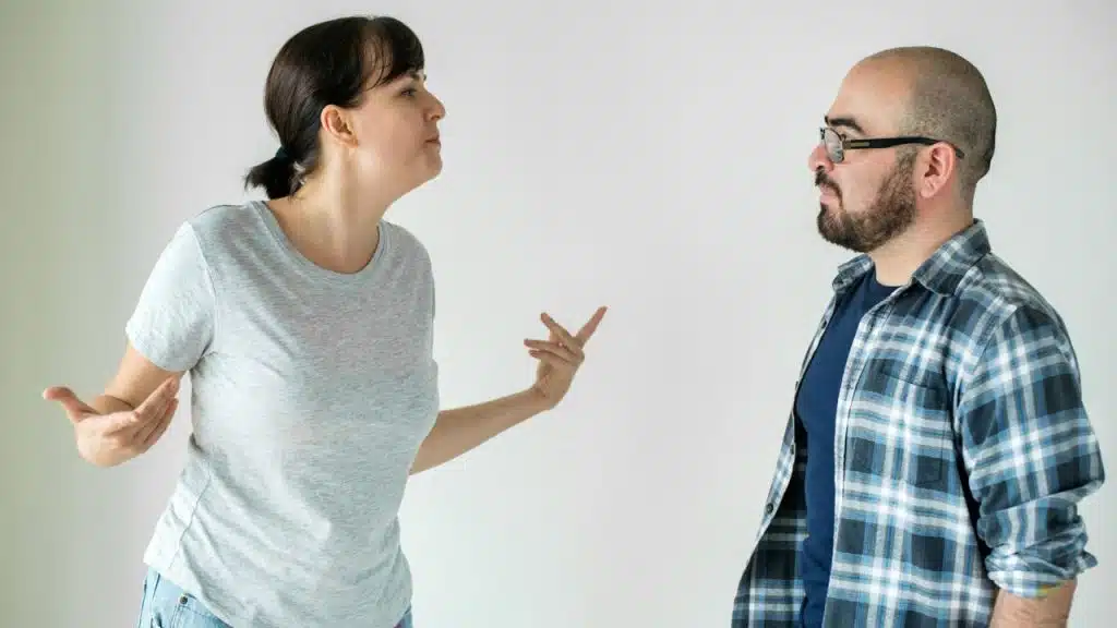 A woman in a grey shirt gestures with both hands while speaking to a bearded man.