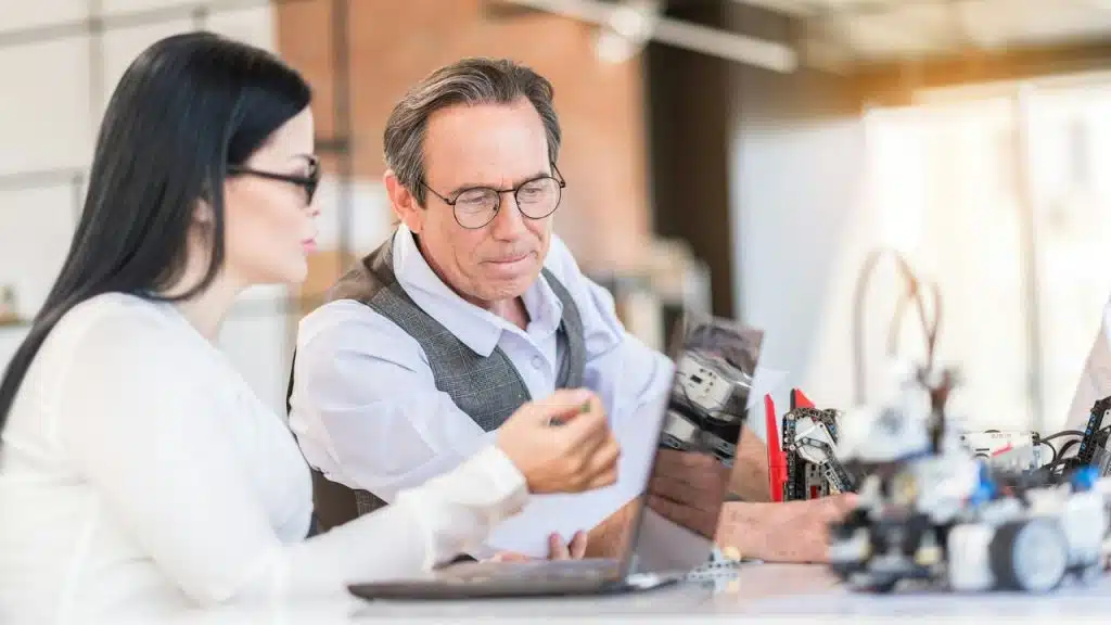 A woman and man wearing glasses look at a laptop screen together in a bright office.