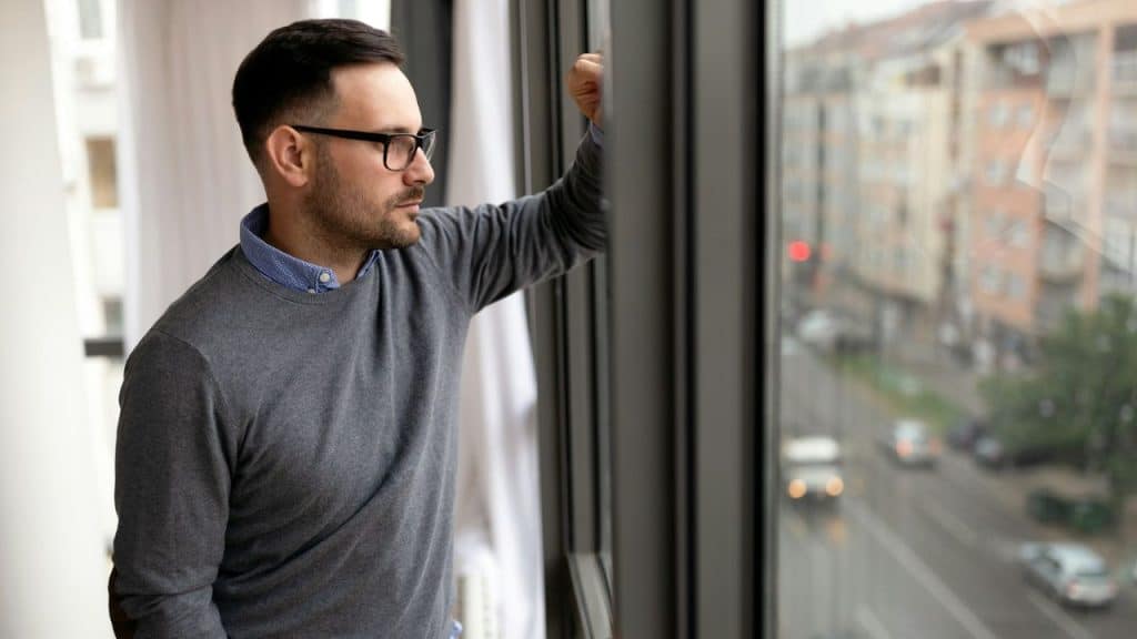 A man wearing glasses and a grey sweater looks out of a large city window.