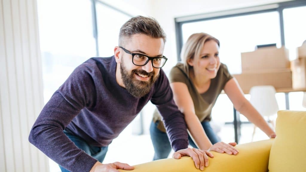 A smiling man with glasses and a woman push a yellow sofa in a bright room.