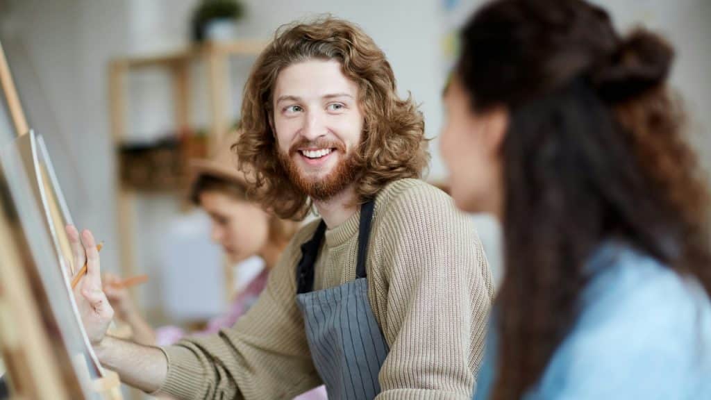 A man with long curly hair and an apron smiles while painting at an easel.