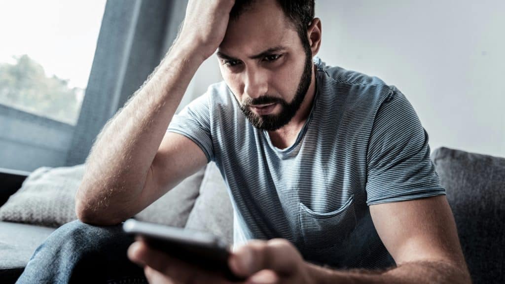 A bearded man sits on a couch holding his head while looking down at a smartphone.