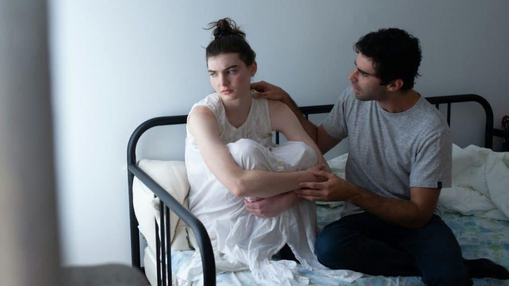 A man sits on a bed reaching toward a woman who is looking away somberly.