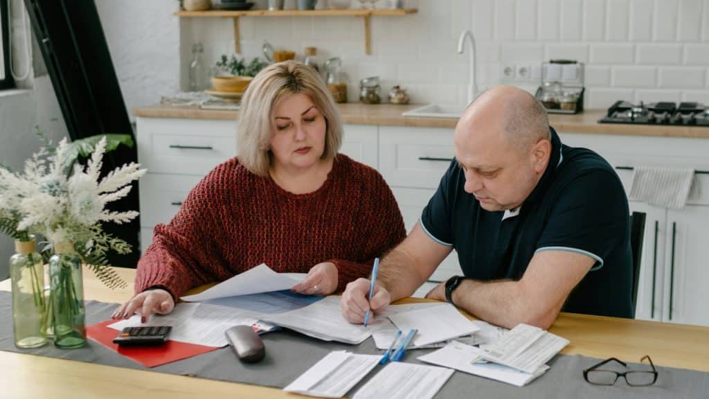 A man and woman sit at a kitchen table surrounded by papers, a calculator, and pens.
