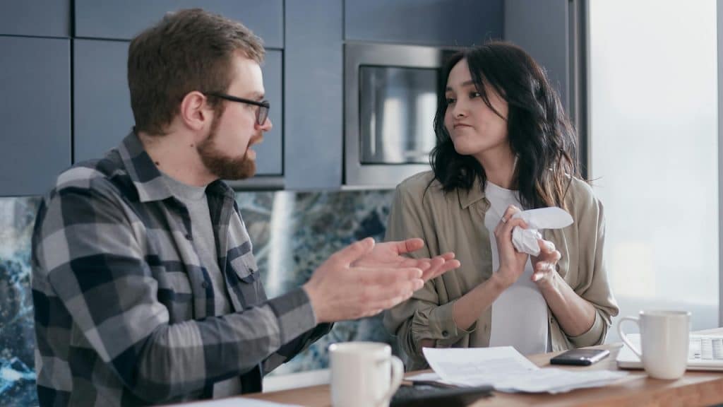 A man gestures toward a woman holding a crumpled piece of paper in a kitchen.