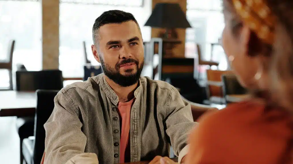 A man with a dark beard sits at a table looking at a person off-camera.