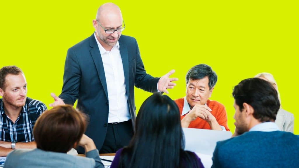 A man in a suit stands gesturing with his hands while addressing a seated group.