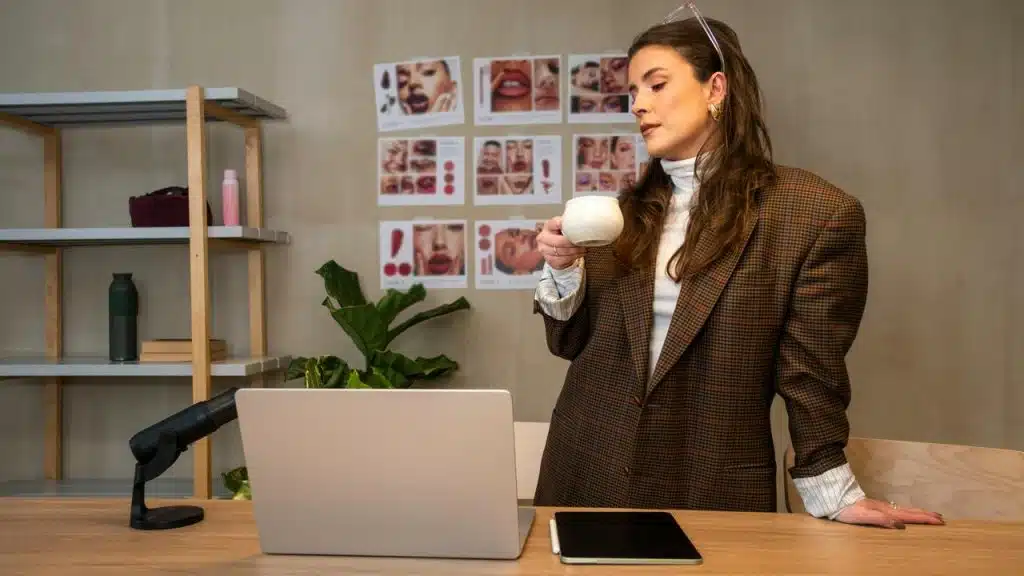 A woman holding a cup while standing at a desk with a laptop in a studio setting.