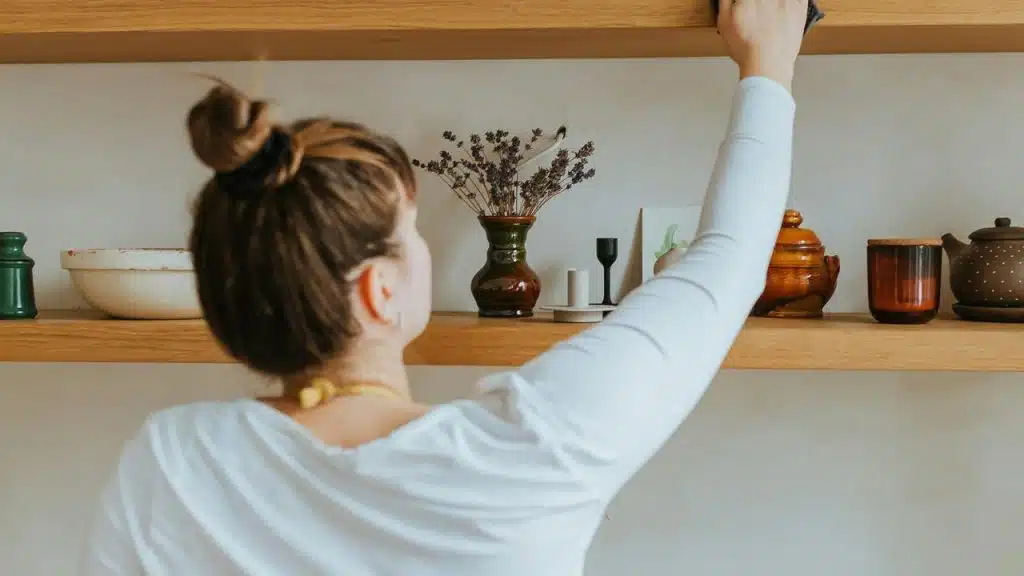 A woman reaches up to clean or arrange items on a wooden shelf.