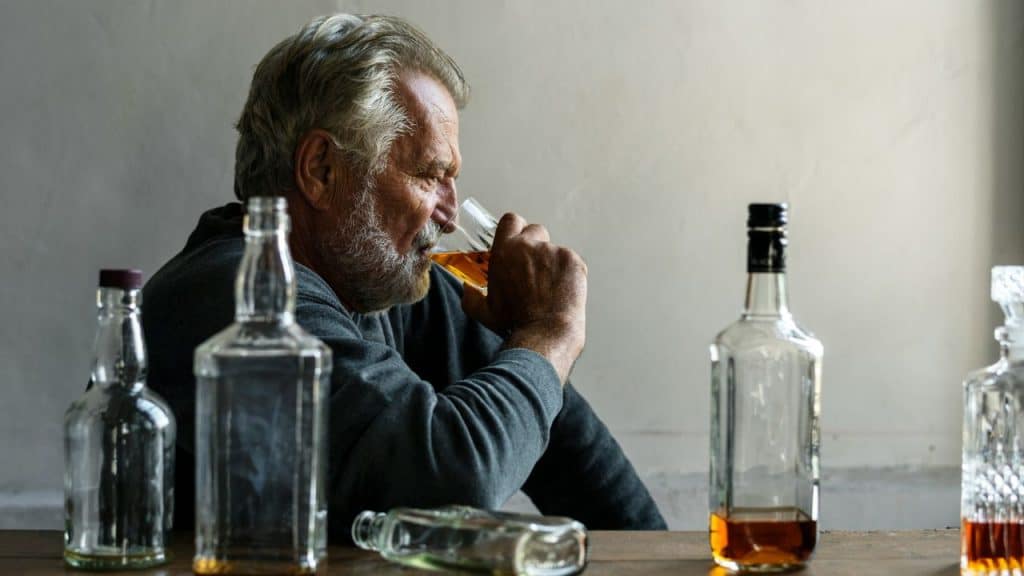 A man with grey hair drinks from a glass surrounded by several glass bottles.