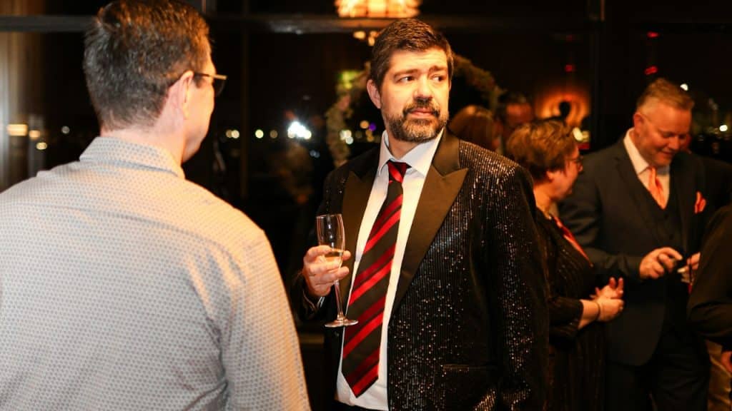 A man in a sequined blazer holds a glass while talking to others at a party.