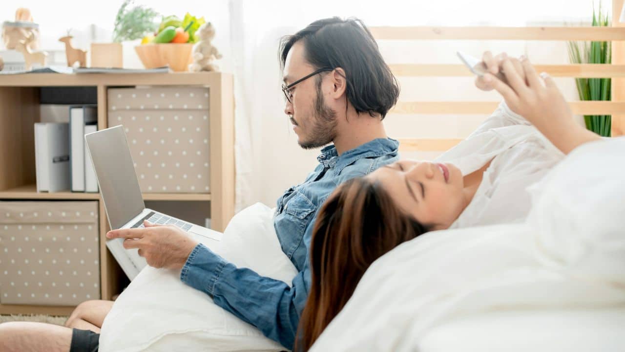 A man uses a laptop in bed while a woman lies beside him using a phone.