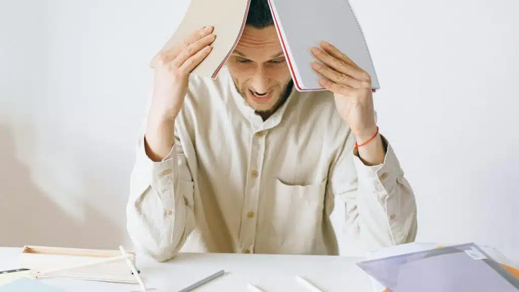 A stressed man holding open notebooks over his head while sitting at a desk.