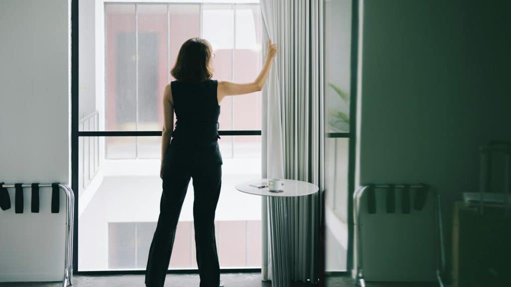 A woman standing by a window, opening a curtain to look outside.