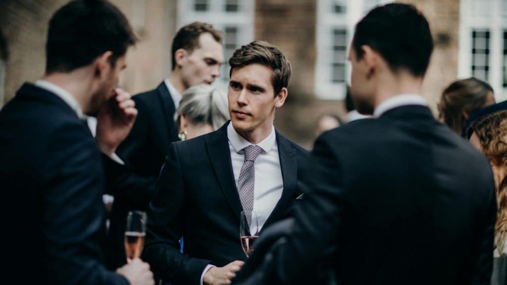 A man in a dark suit and patterned tie stands at a gathering holding a glass.