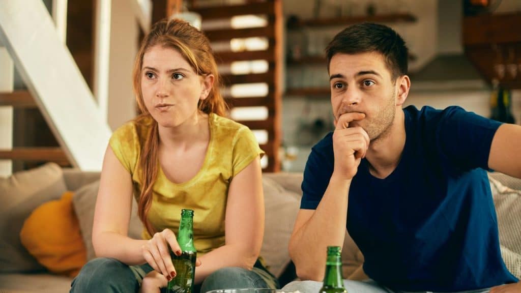 A man and woman sit on a couch, holding green bottles and looking forward intently.
