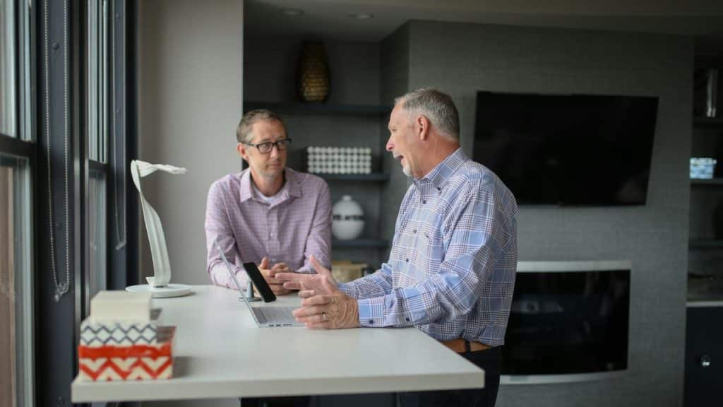 Two men in plaid shirts have a discussion while standing at a high desk.