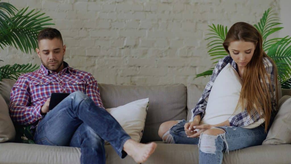 A man uses a tablet on a sofa next to a woman holding a pillow.