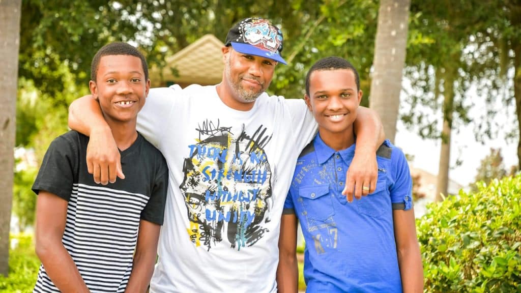 A man standing with his two sons and resting his arms on their shoulders.