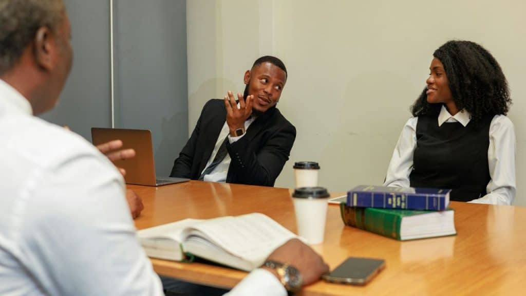 Three professionals sit at a conference table with books, laptops, and coffee cups.