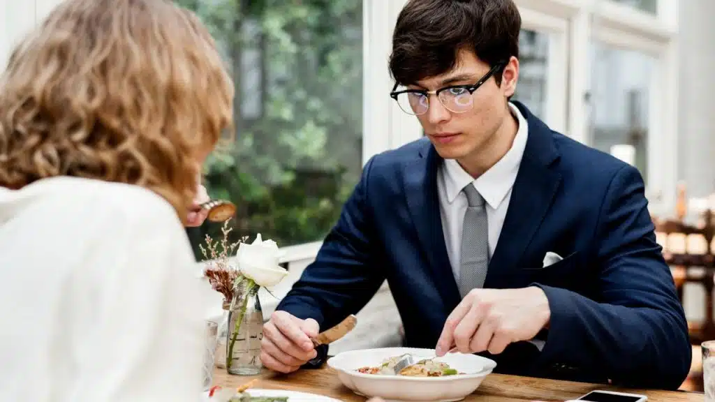 A man in a suit and glasses eats with a woman at a table.