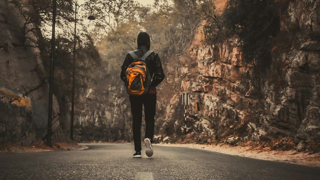 A man wearing a backpack walking on a road surrounded by mountains.