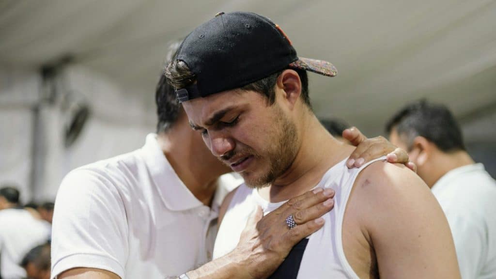 A man wearing a baseball cap cries while another person places a hand on him.