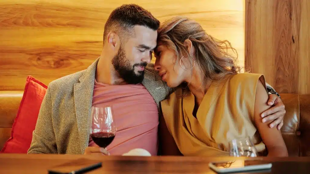 A couple sitting together at a table, resting their heads against one another with wine.