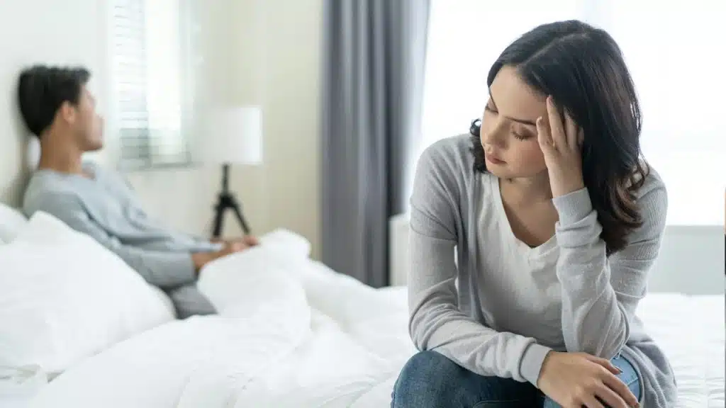 An upset woman sitting on the edge of a bed while her husband lies in the bed behind her.