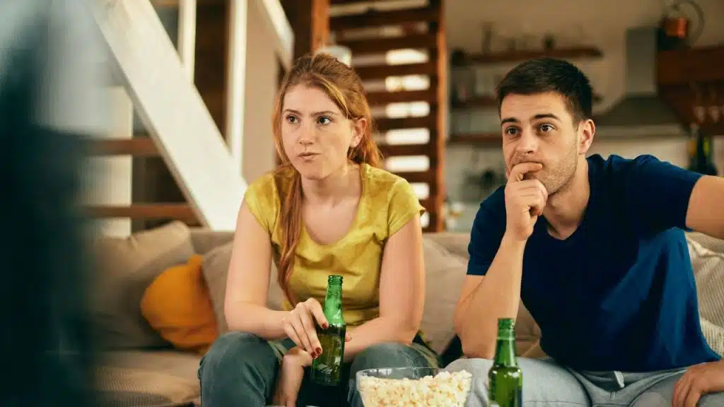A woman and man sit on a couch with popcorn and green glass bottles.
