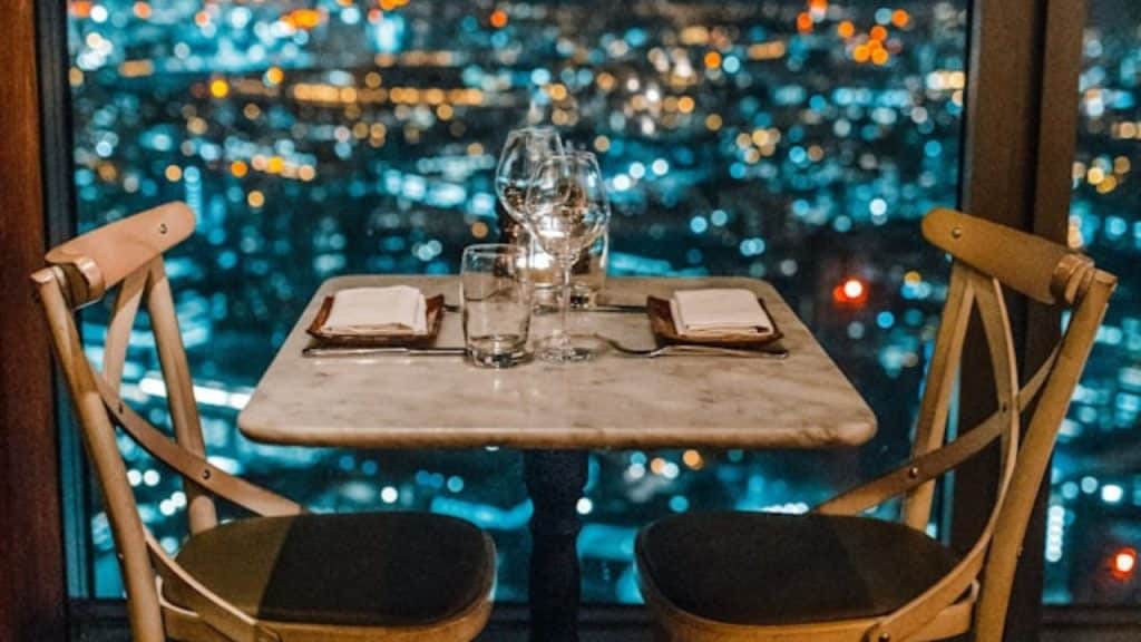 A set dinner table for two stands empty before a blurred nighttime city skyline window.